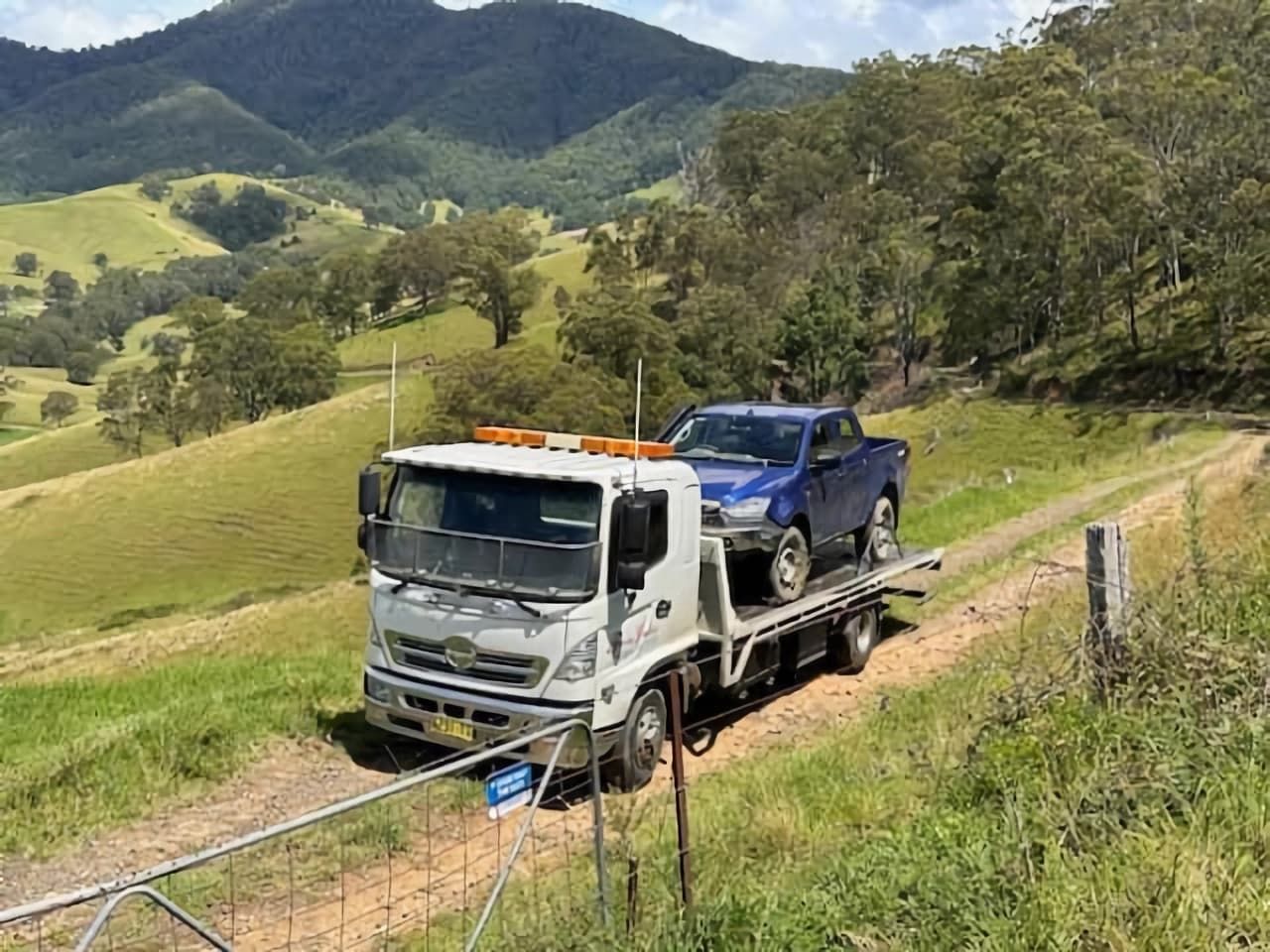 A Tow Truck Is Carrying a Blue Truck Down a Dirt Road — Twin Lakes Towing & Recovery in Hamlyn Terrace, NSW