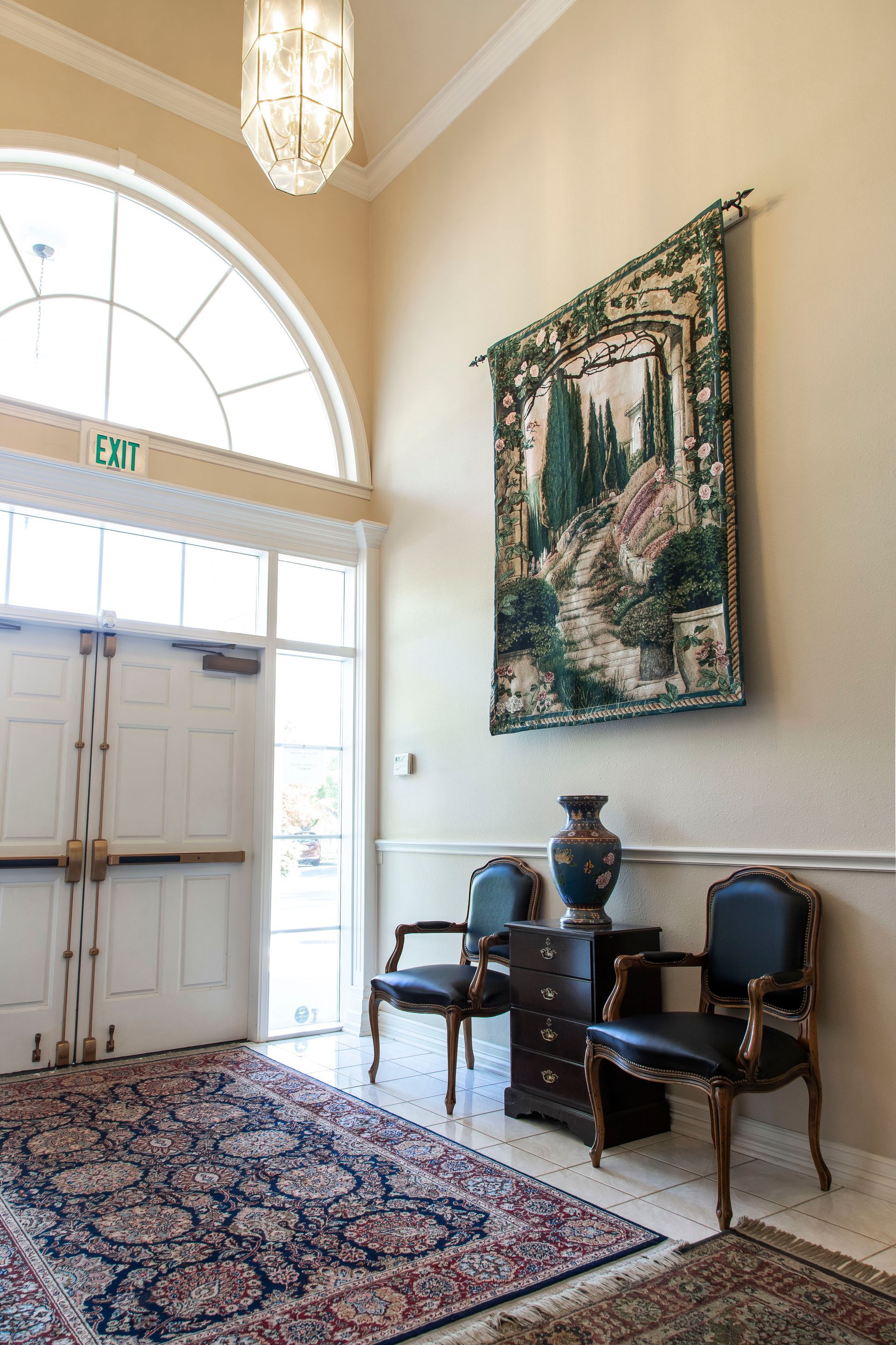 Entryway with arched door, tapestry, chairs, rug, and chandelier. Beige walls, wood accents, and ornate decor.