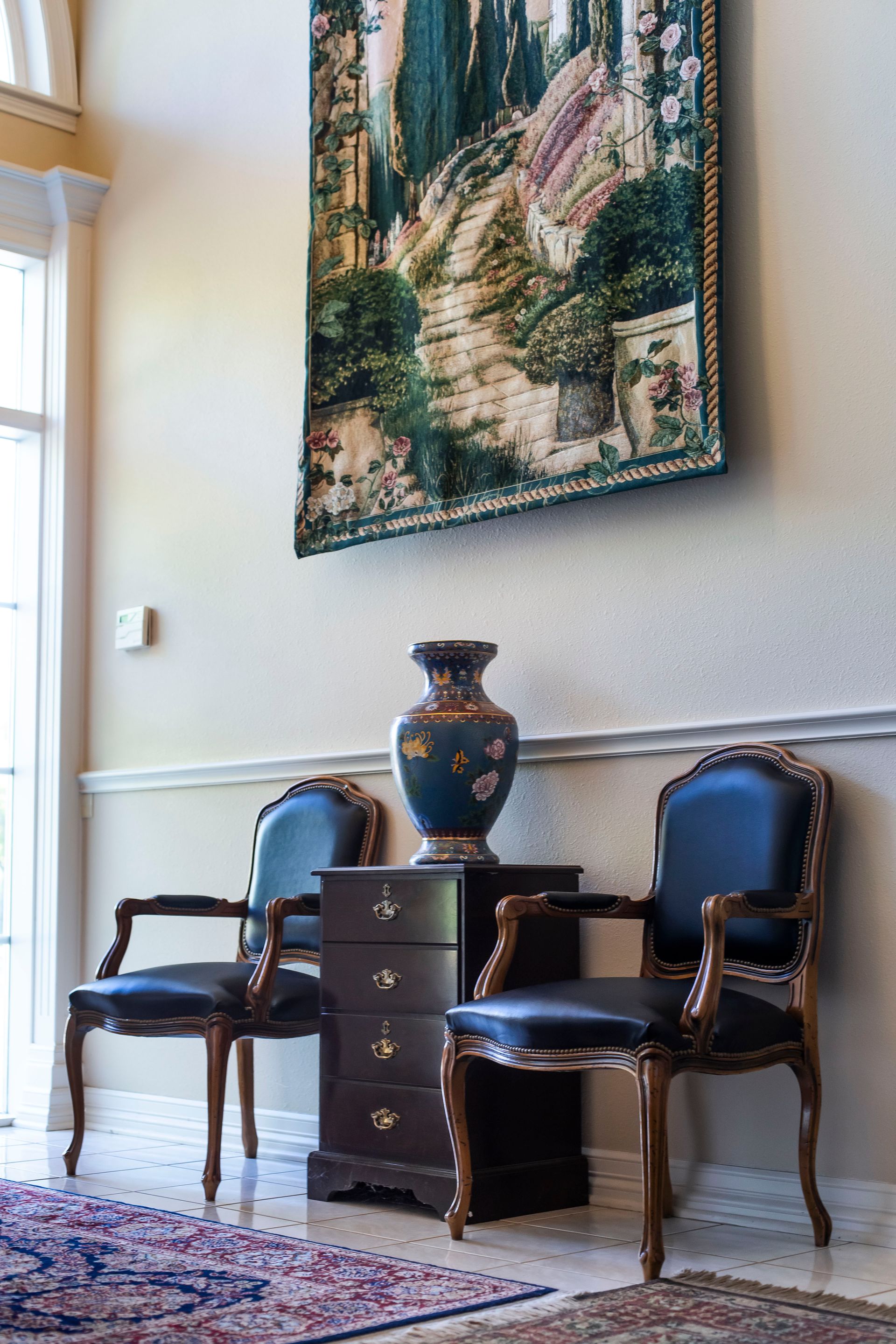 Two dark blue chairs flanking a small wooden cabinet holding a decorative vase, beneath a landscape tapestry.