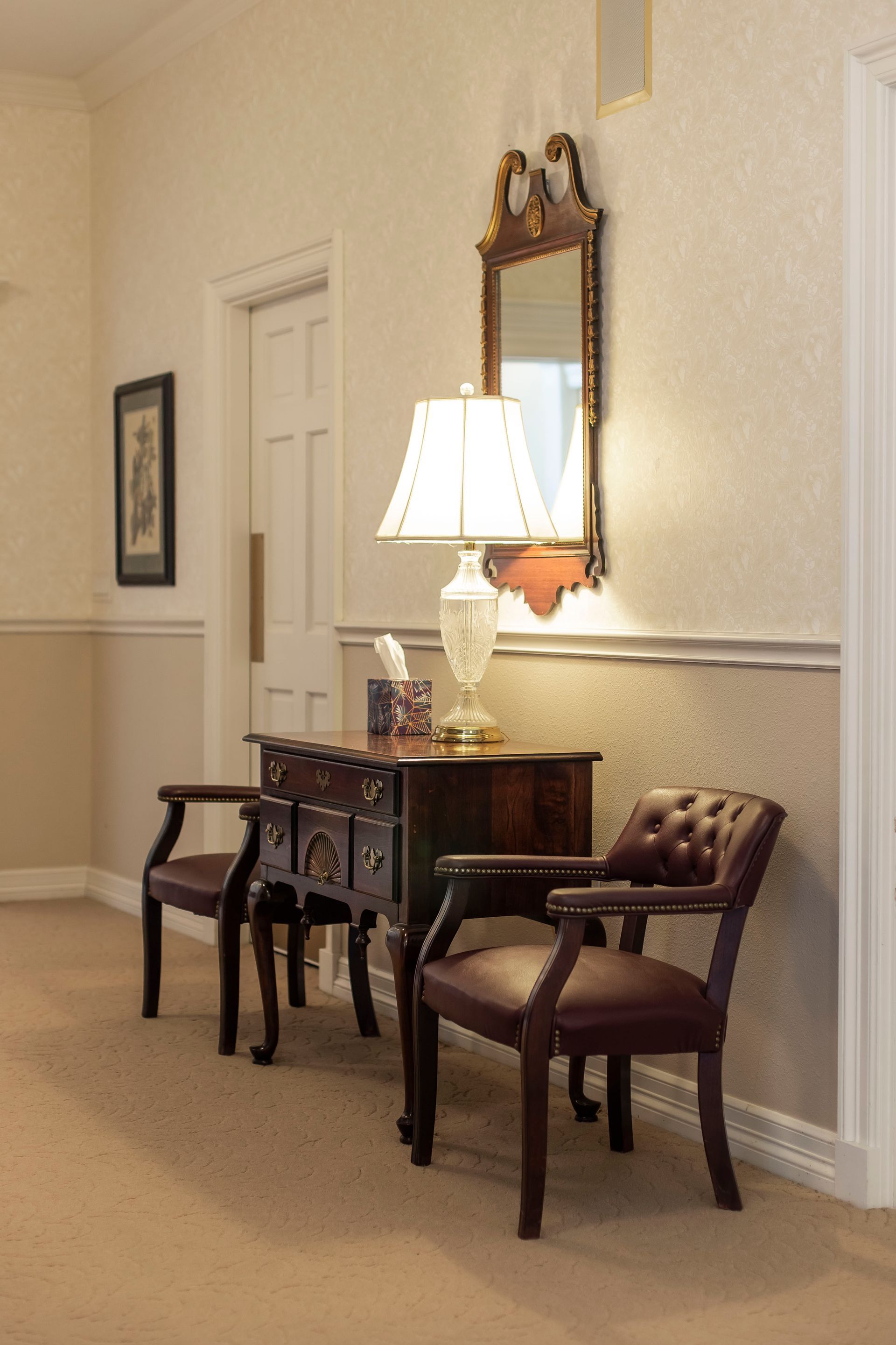 Entryway with ornate mirror, lamp, small wooden table, and two chairs. Beige and brown tones.