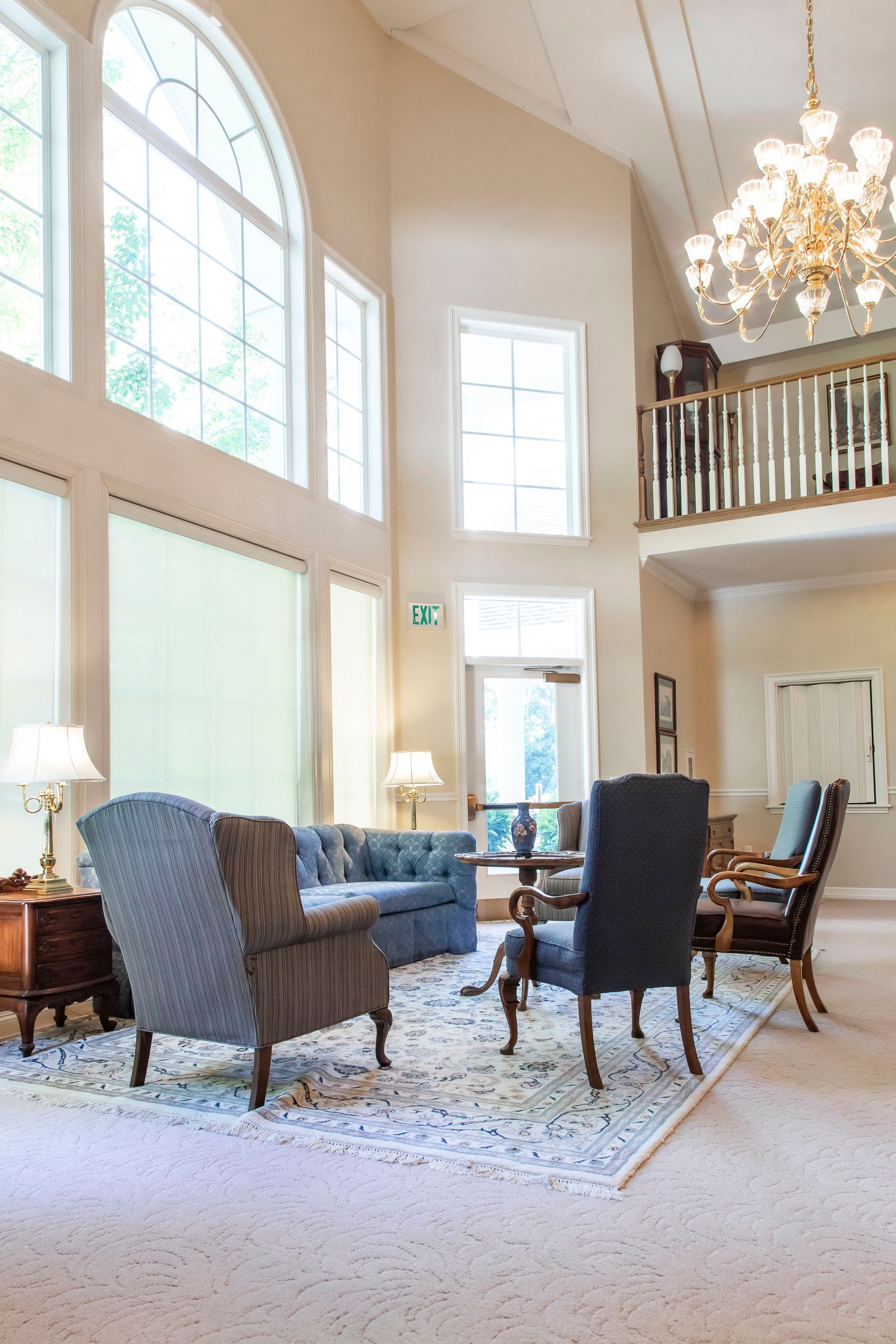 Brightly lit lobby with seating area; large windows, high ceiling, chandelier, and a balcony above.