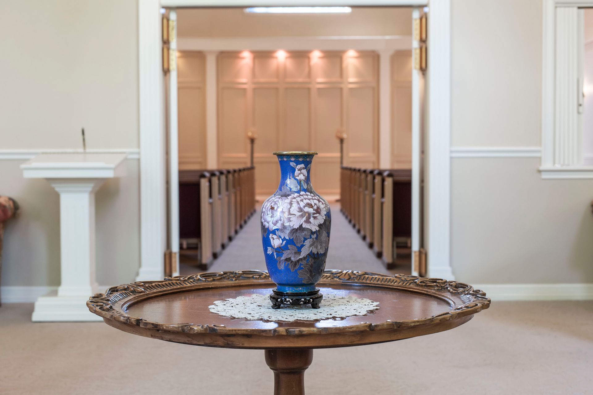 Blue vase on a round table, centered in a room with a doorway leading to rows of pews.