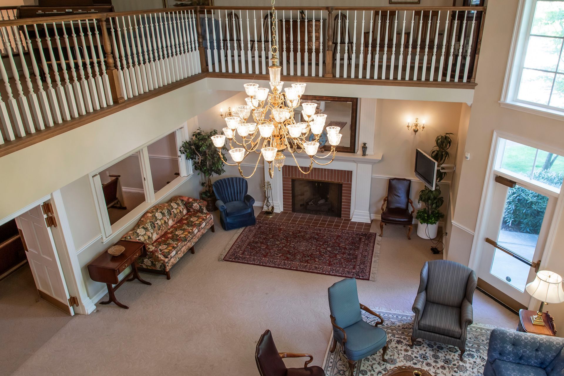 Spacious living room with a chandelier, fireplace, and seating area. Balcony overhead, natural light streams in.