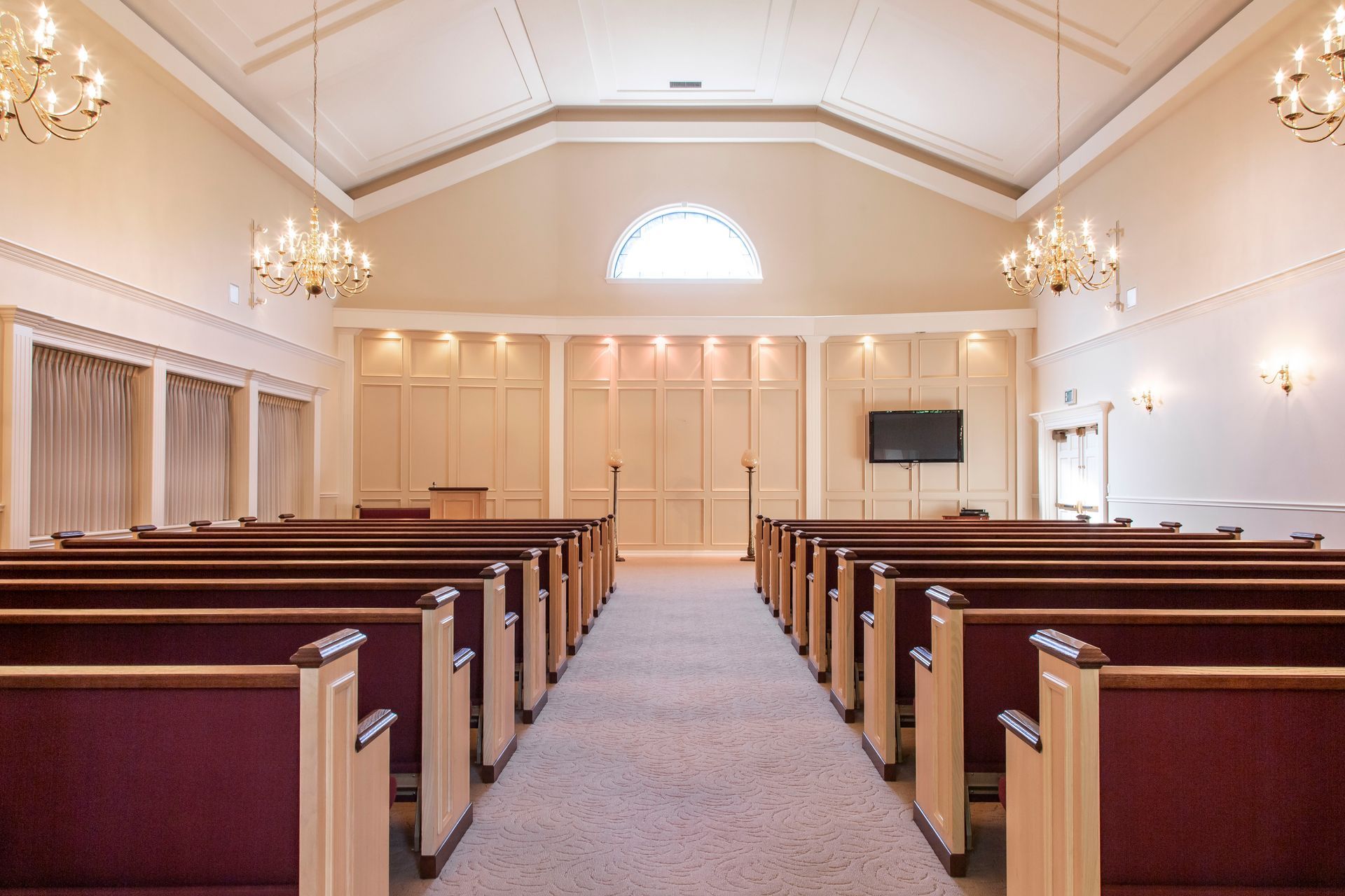 Interior of a beige chapel with rows of wooden pews and burgundy seats. There's a pulpit and a TV screen.