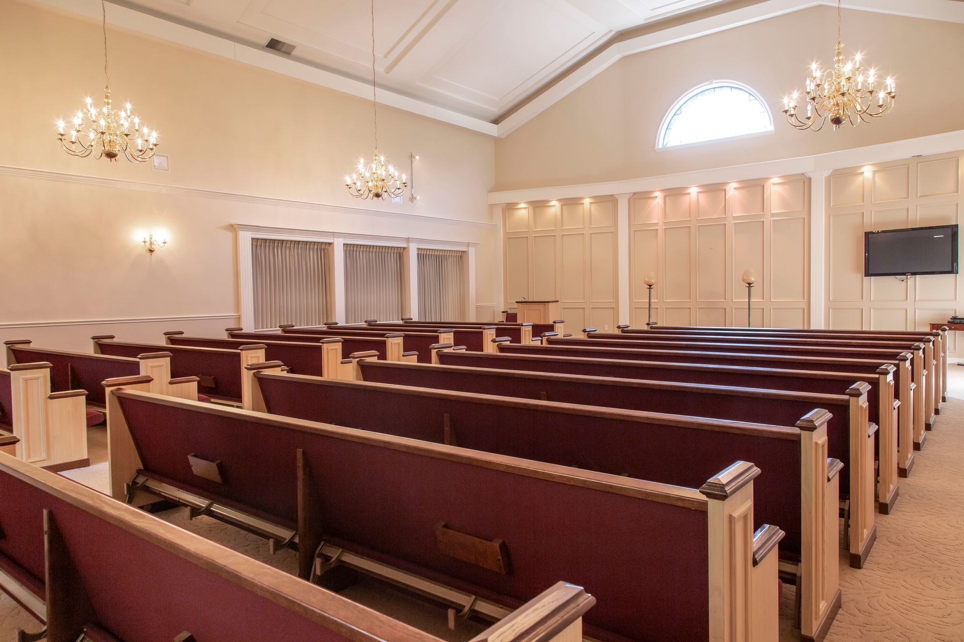 Empty chapel with rows of pews, chandeliers, and stage with a TV.
