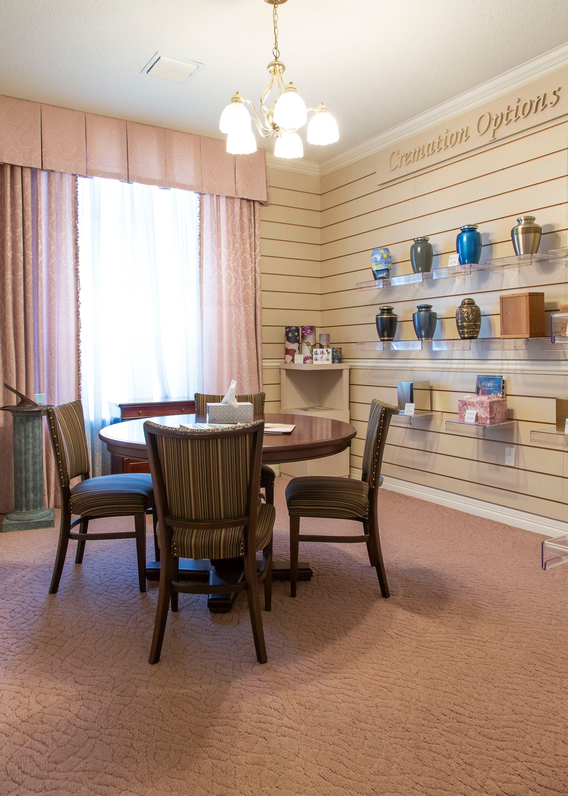 A room with a round table and chairs, display of urns, and a window with pink curtains.