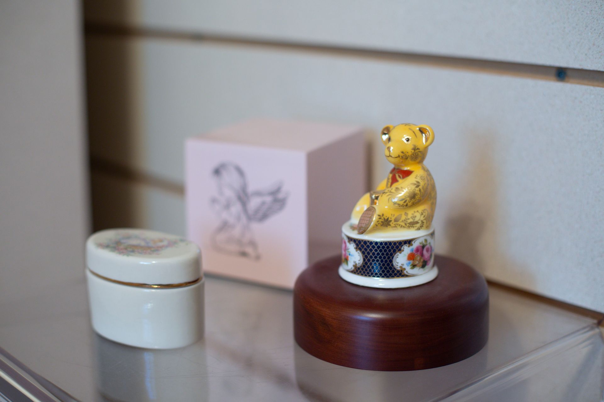 Yellow teddy bear figurine on a wooden stand, with a small box and container on a shelf.