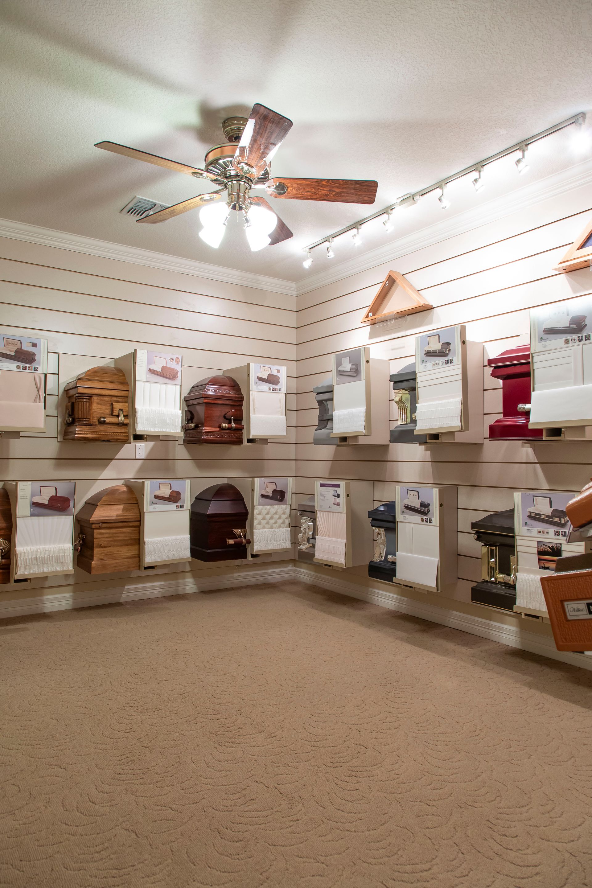 Interior of a showroom displaying various caskets, with a ceiling fan overhead and neutral carpet.