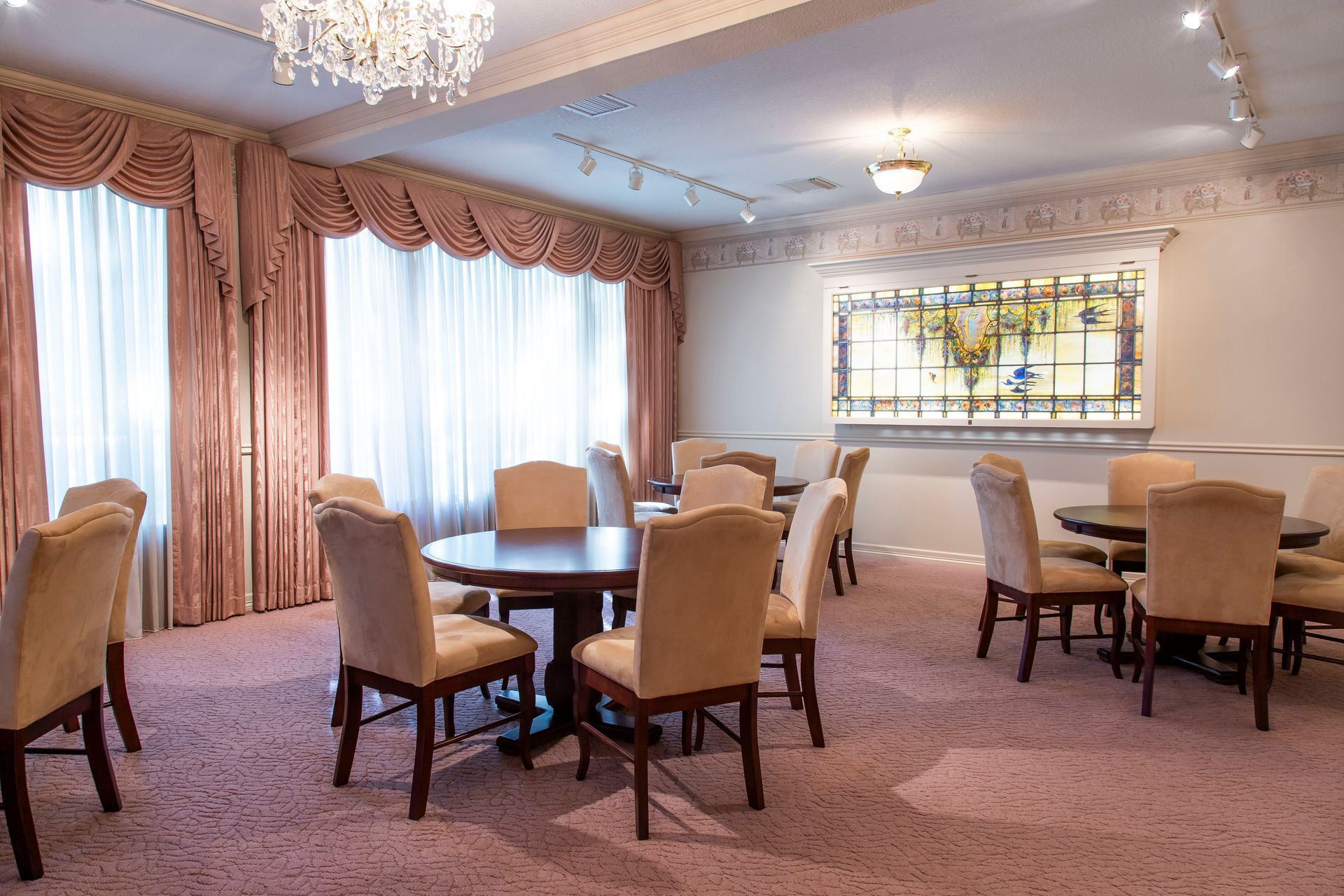A room with round tables, chairs, and pink carpet, drapes, and a chandelier.