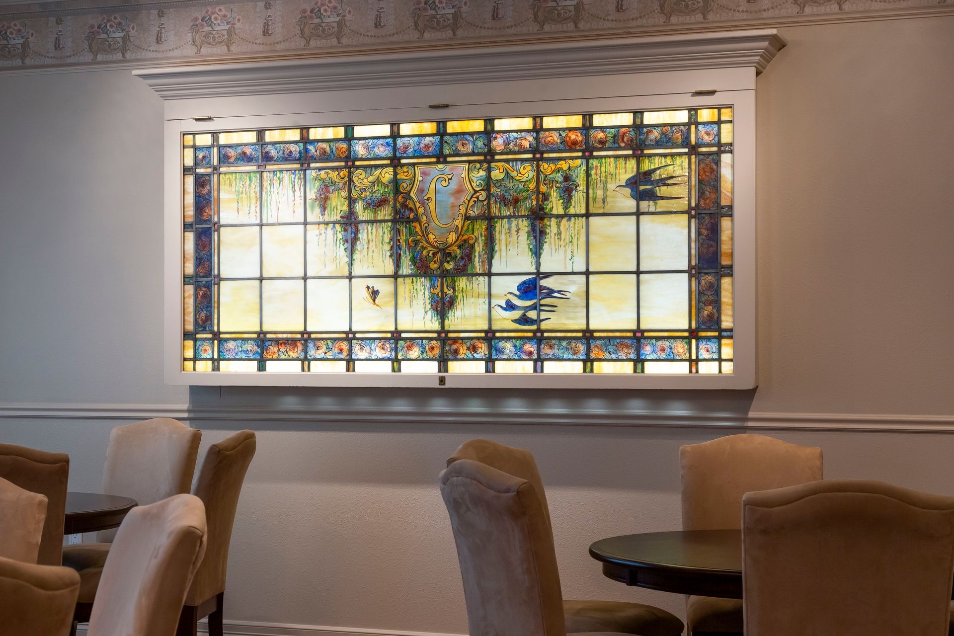 Stained glass window depicting a pond scene with birds; above chairs and tables in a dining area.