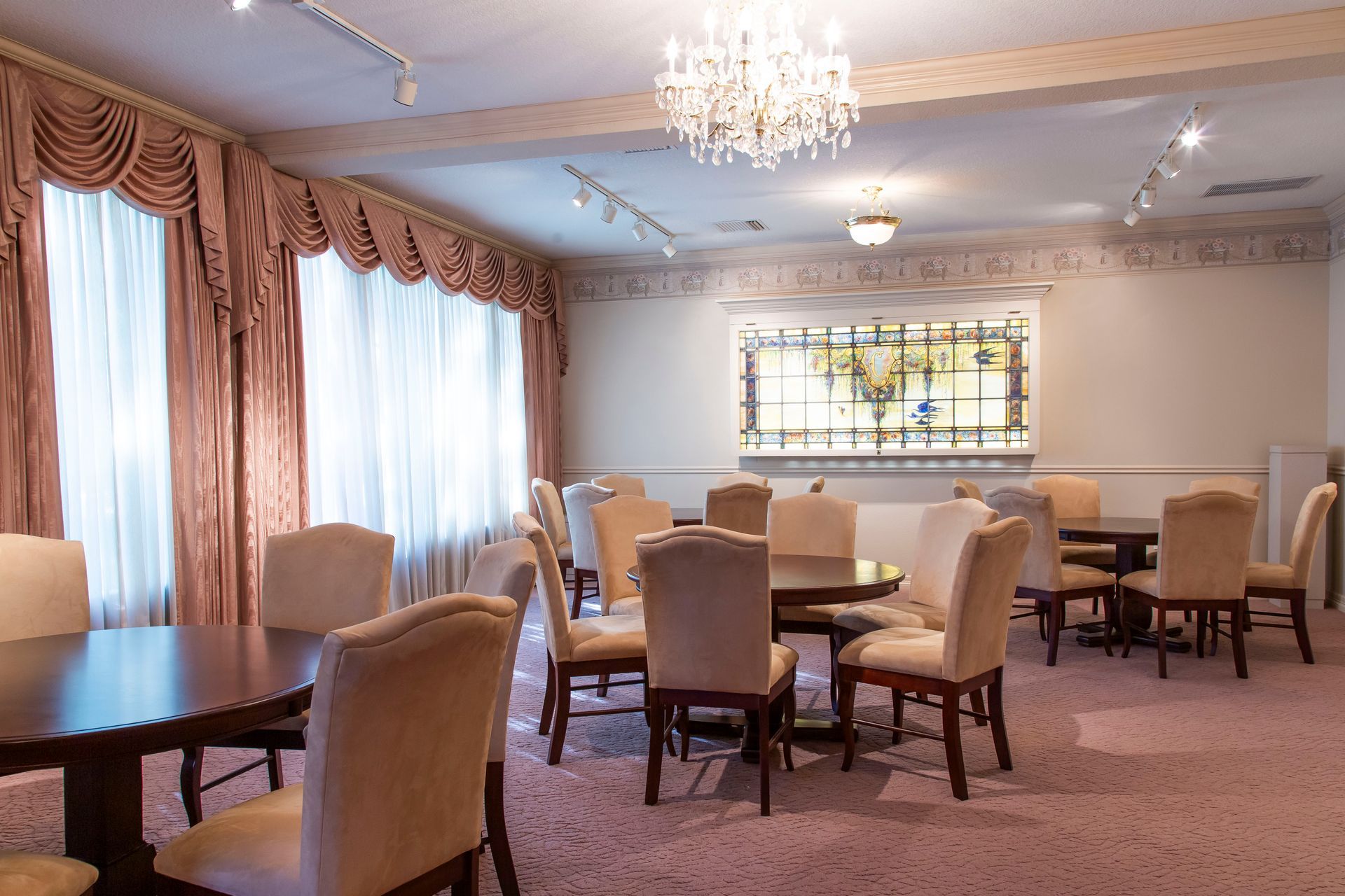 Interior of a room with round tables, chairs, and large window with pink curtains. Chandelier hangs from ceiling.