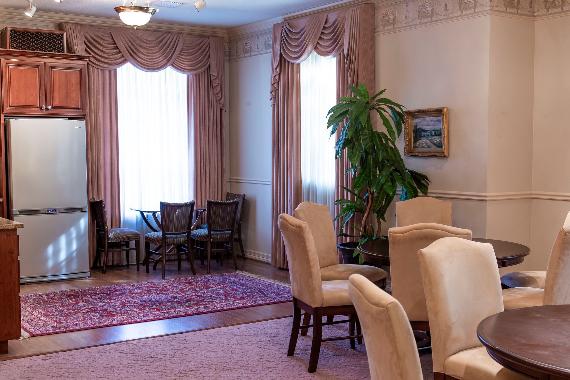 Elegant dining room with pink curtains, rug, and chairs. Refrigerator and cabinets on the left.