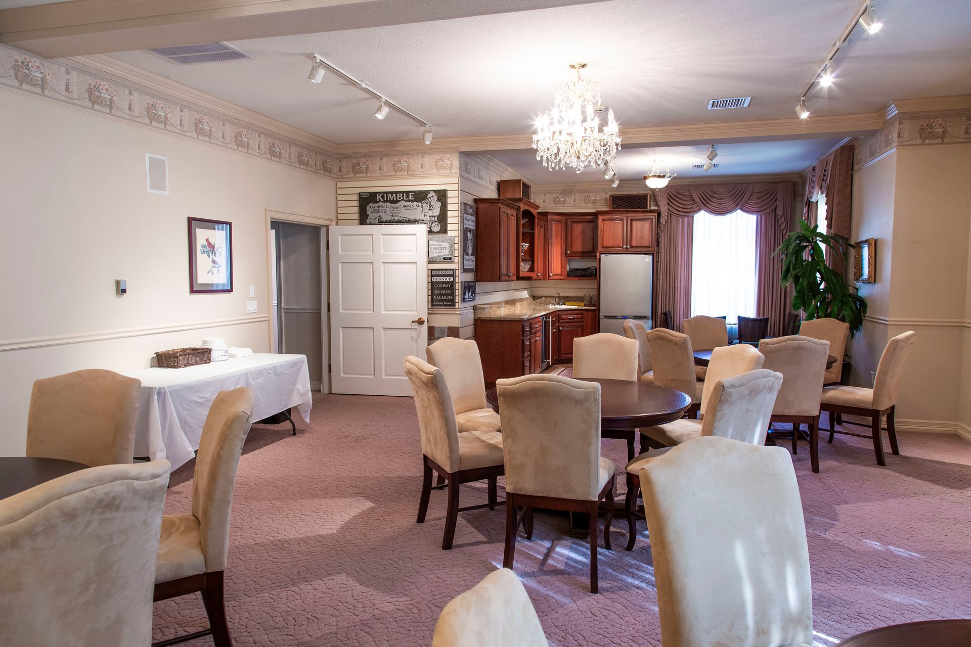 Interior of a room with chairs around round tables, a small kitchen, and a chandelier.