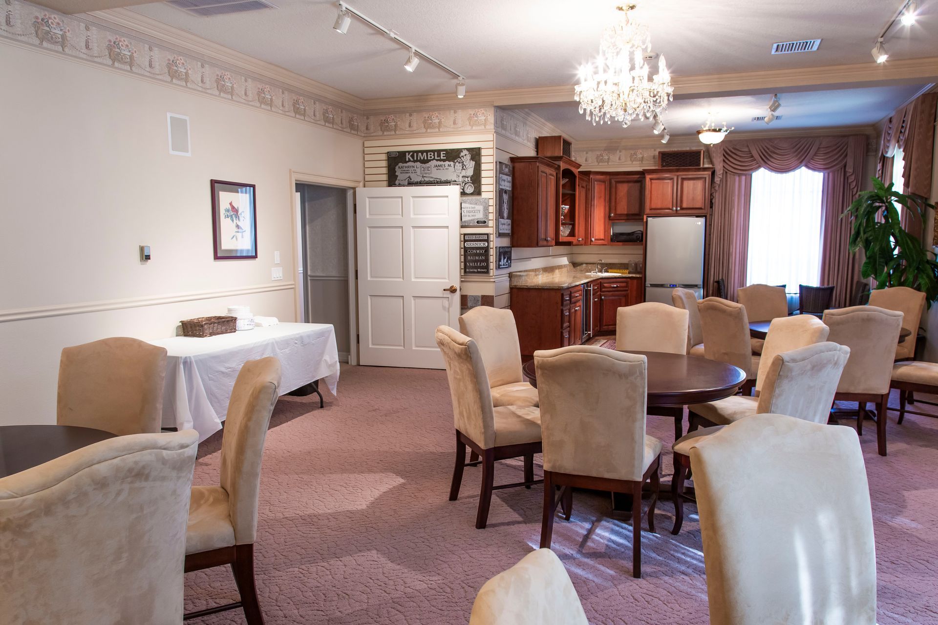 Interior with tables, chairs, and a small kitchen. Pink carpet, light walls, and a chandelier.