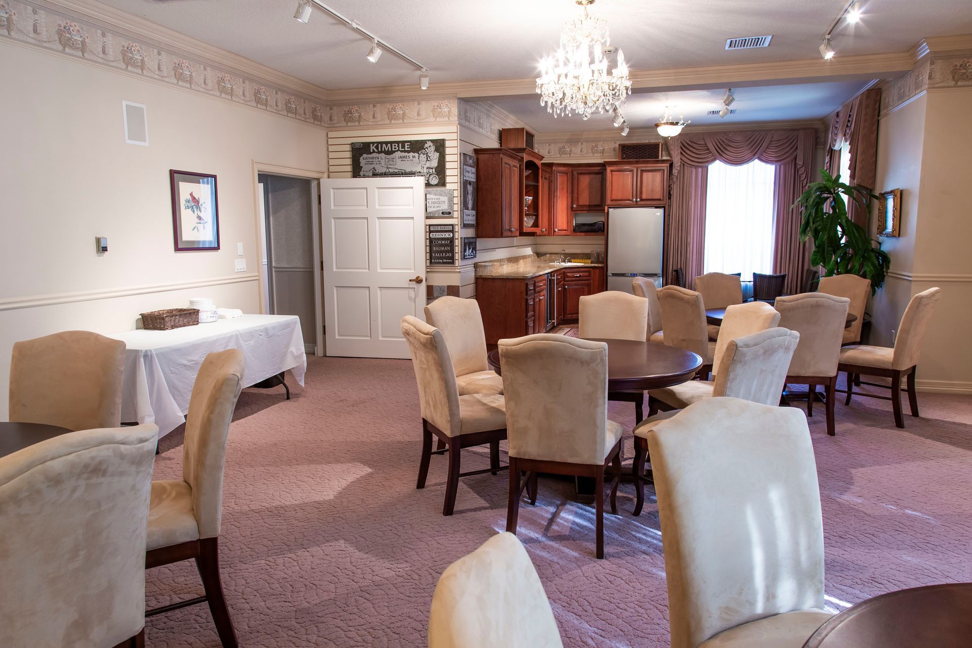Interior of a room with chairs around round tables, small kitchen, chandelier, and a table with a white tablecloth.