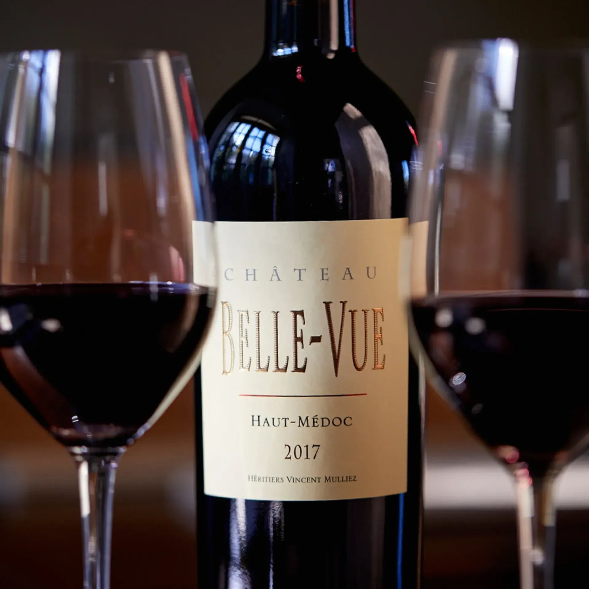 Bottle of Chateau Belle-Vue Haut-Médoc 2017 red wine with two filled wine glasses in a dimly lit setting.