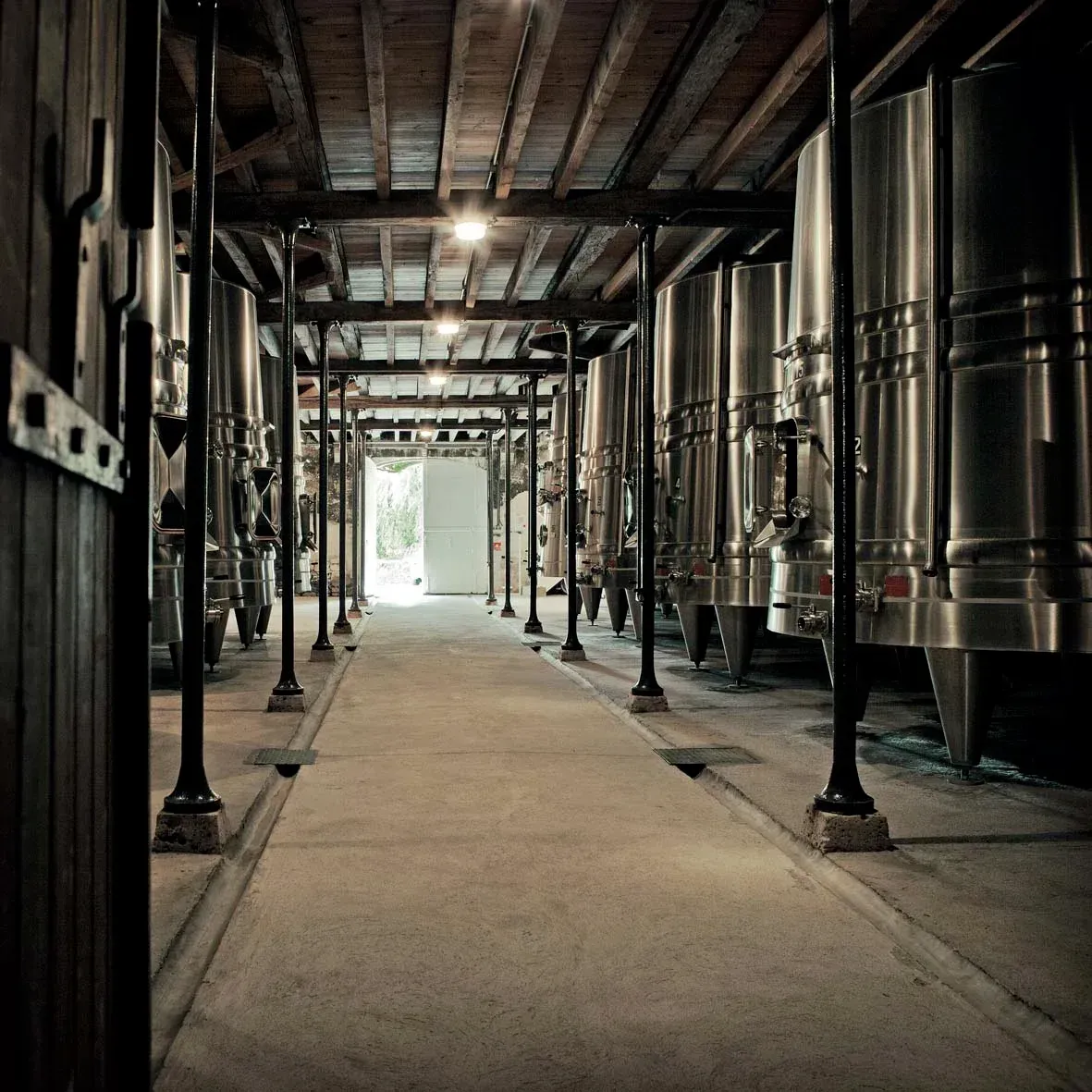 Stainless steel tanks line a concrete floor with wooden beams overhead, leading to a bright doorway.