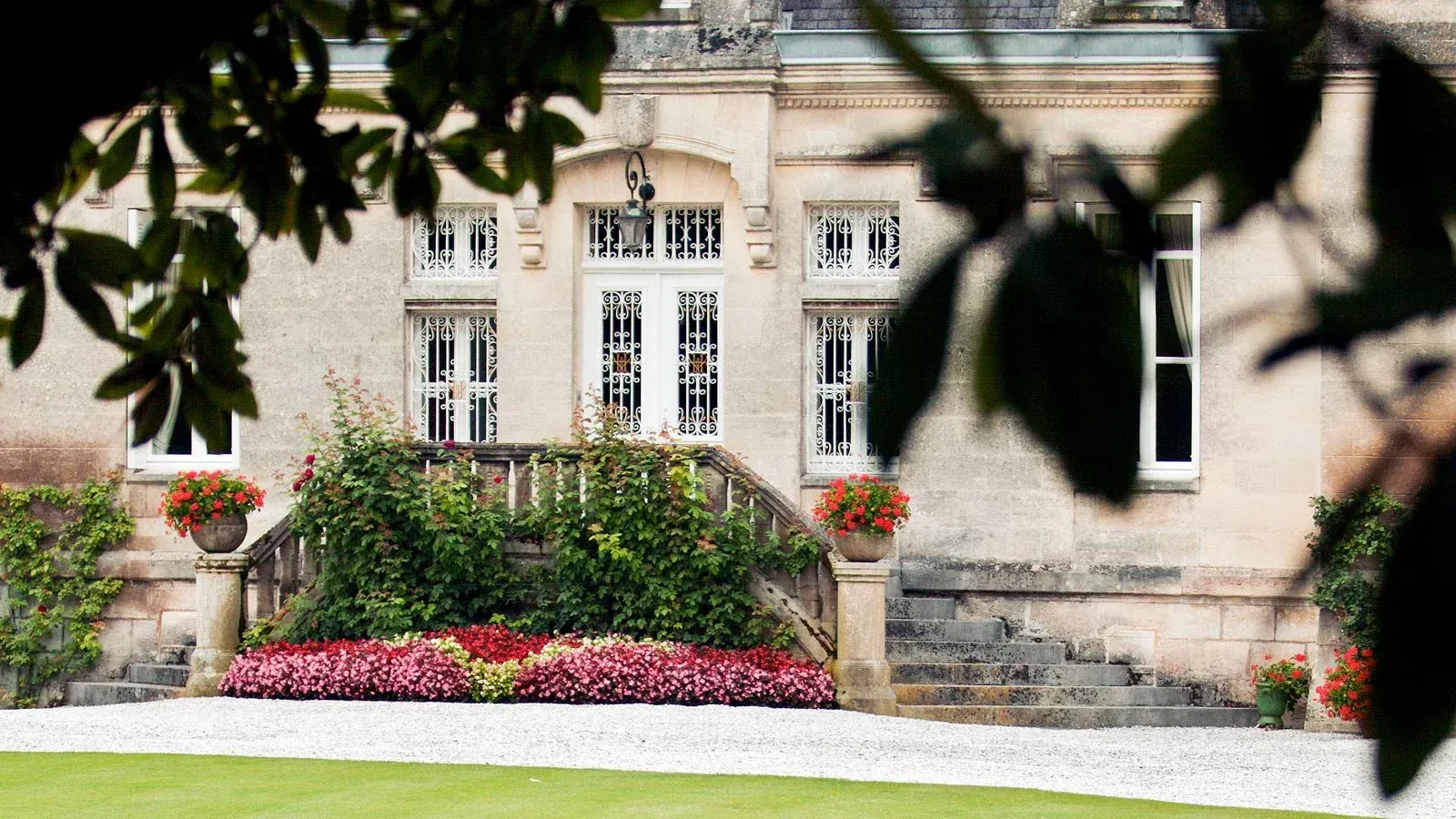 Stone building entrance with steps, flowerbeds at Château Bellegrave, and a wrought-iron door. 