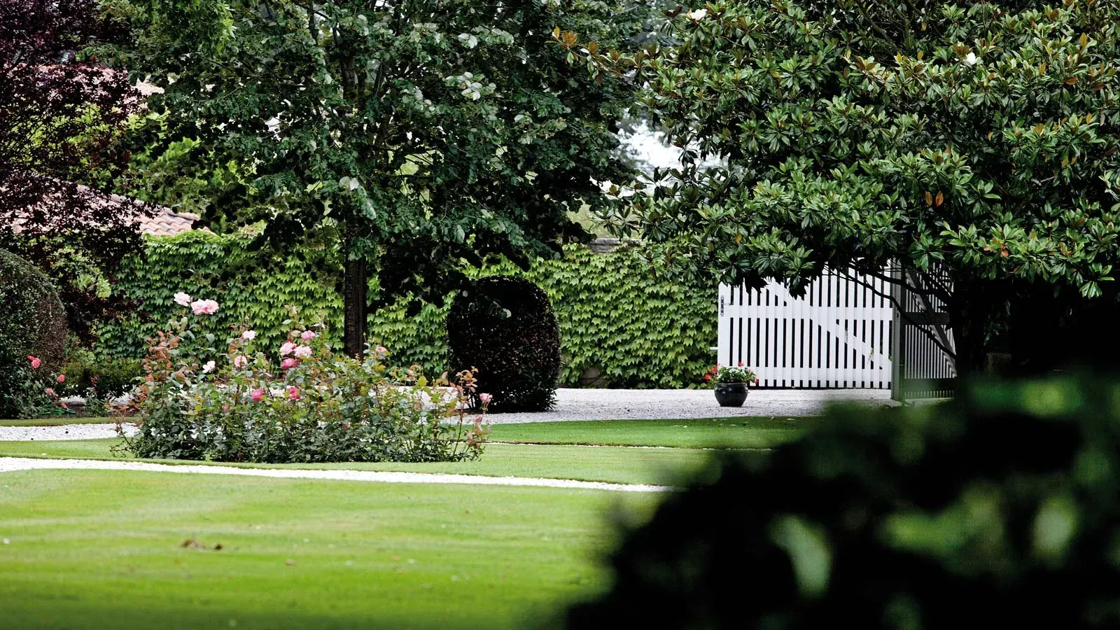 Lush green lawn at Château Bellegrave, leading to a white picket gate in a garden surrounded by trees and flowers.