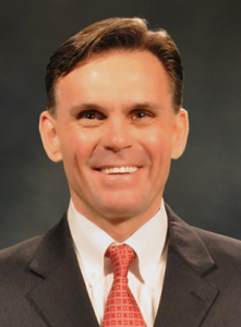 A headshot of a person wearing a dark suit, white shirt, and patterned red tie, smiling against a dark background.