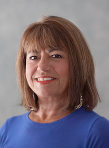 A smiling person with shoulder-length brown hair, wearing a blue shirt and hoop earrings against a gray background.