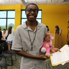 Girl with brown hair, smiling, wearing a hairnet, gloves, and a green shirt, leaning on a table.