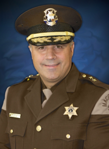 A smiling law enforcement officer in a brown uniform with a peaked cap and sheriff’s star badge against a blue background.