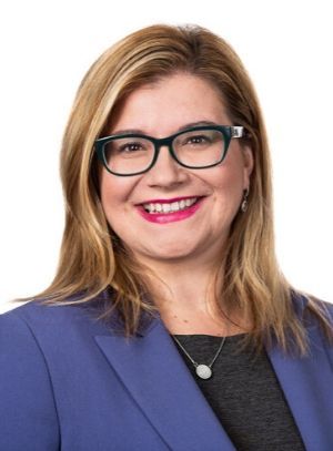 Headshot of a smiling person wearing glasses, a blue blazer, and a necklace against a white background.