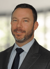A professional headshot of a person with a short beard, wearing a gray suit, a light patterned shirt, and a dark tie.