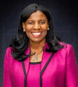 A smiling person wearing a magenta blazer and a silver pendant necklace against a dark background.