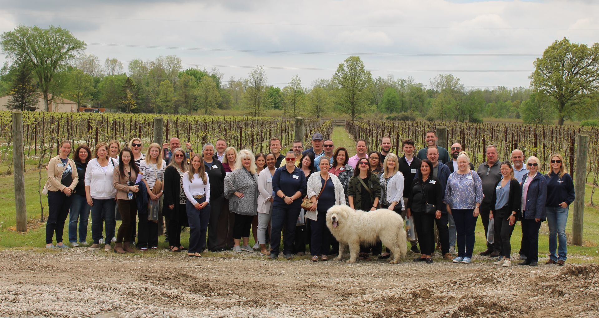 A group of people standing together in a vineyard, with a large, fluffy white dog standing in the front row.