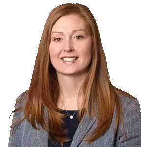 A professional portrait of a person with long reddish-brown hair wearing a blue blazer and a simple pendant necklace.
