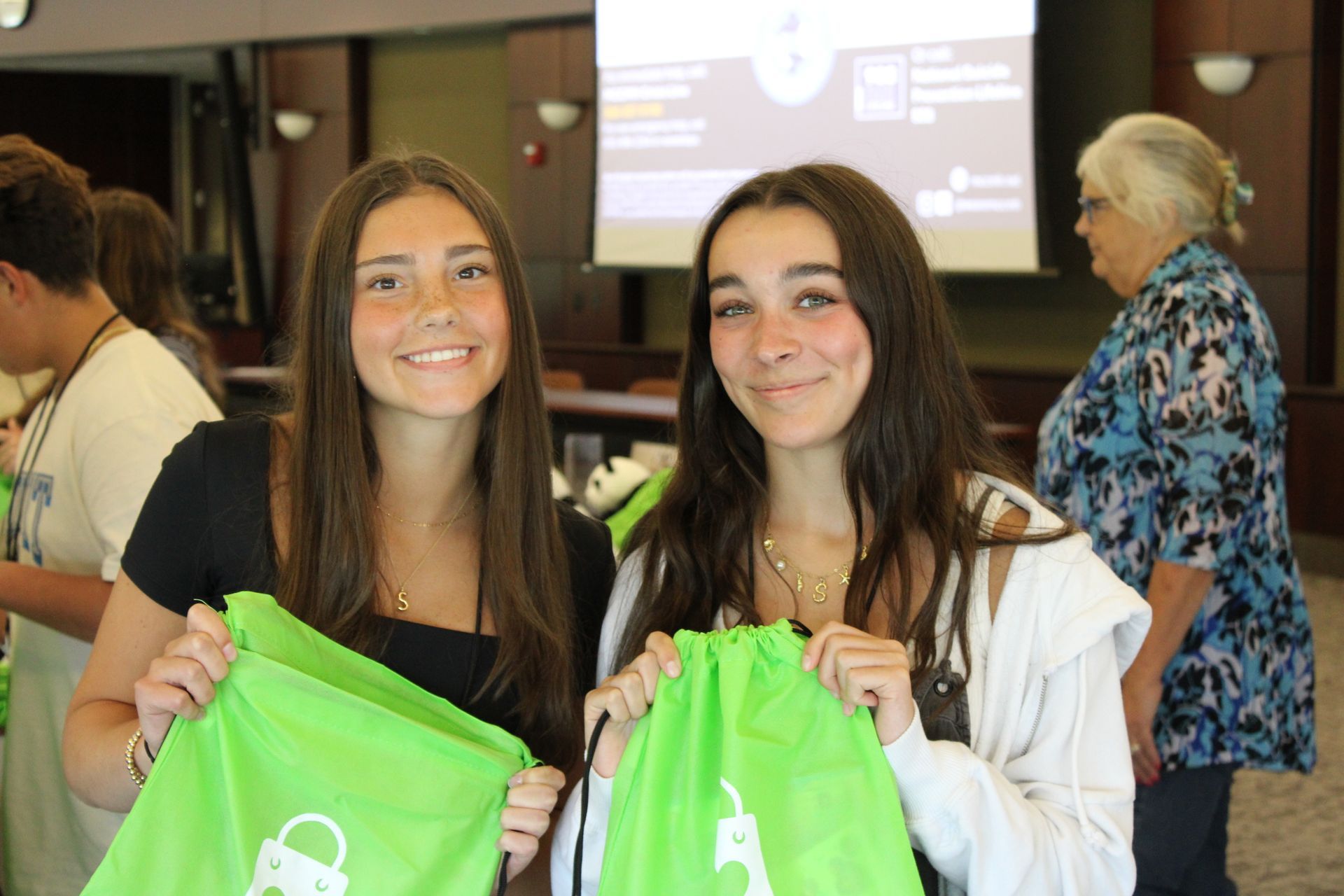Two people smiling, holding bright green drawstring bags in an indoor space with a projection screen in the background.