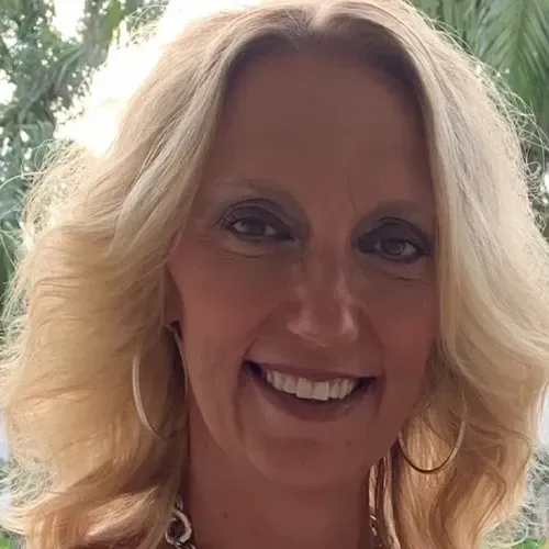 A person with shoulder-length blonde hair and hoop earrings smiling at the camera against a blurred outdoor background.