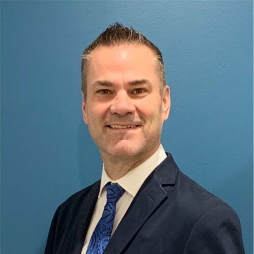 A professional headshot of a person wearing a navy blue blazer, white shirt, and blue patterned tie against a blue wall.