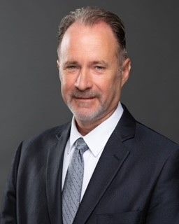 A professional headshot of a man with light brown hair and a short beard, wearing a dark suit, white shirt, and gray tie.