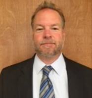 A person in a black suit, white shirt, and blue-and-gray striped tie, standing against a plain brown wood background.