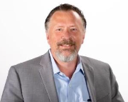 A smiling professional with a goatee wears a light blue collared shirt and gray blazer against a white background.
