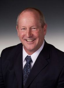 A professional headshot of a smiling person wearing a dark blue suit, white collared shirt, and a patterned blue tie.