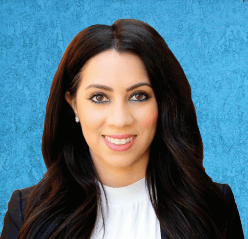 A smiling person with long, dark hair, wearing a white shirt and dark blazer against a textured blue background.