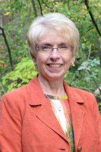 A smiling person wears an orange blazer over a patterned top, set against a blurred green, leafy outdoor background.