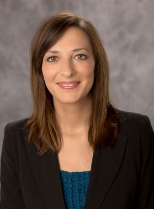 A woman with shoulder-length brown hair, wearing a black blazer and teal top, smiles against a grey mottled background.