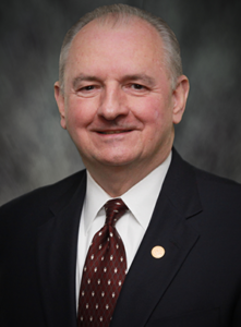 A smiling person in a dark suit, white collared shirt, and patterned burgundy tie against a blurred grey background.
