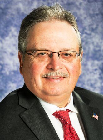 Headshot of a smiling person wearing glasses, a dark suit, and a red tie against a blue textured background.