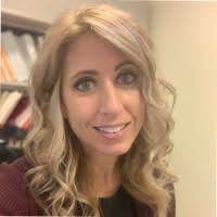 A person with blonde, wavy hair smiles slightly while posing for a headshot in an office setting.