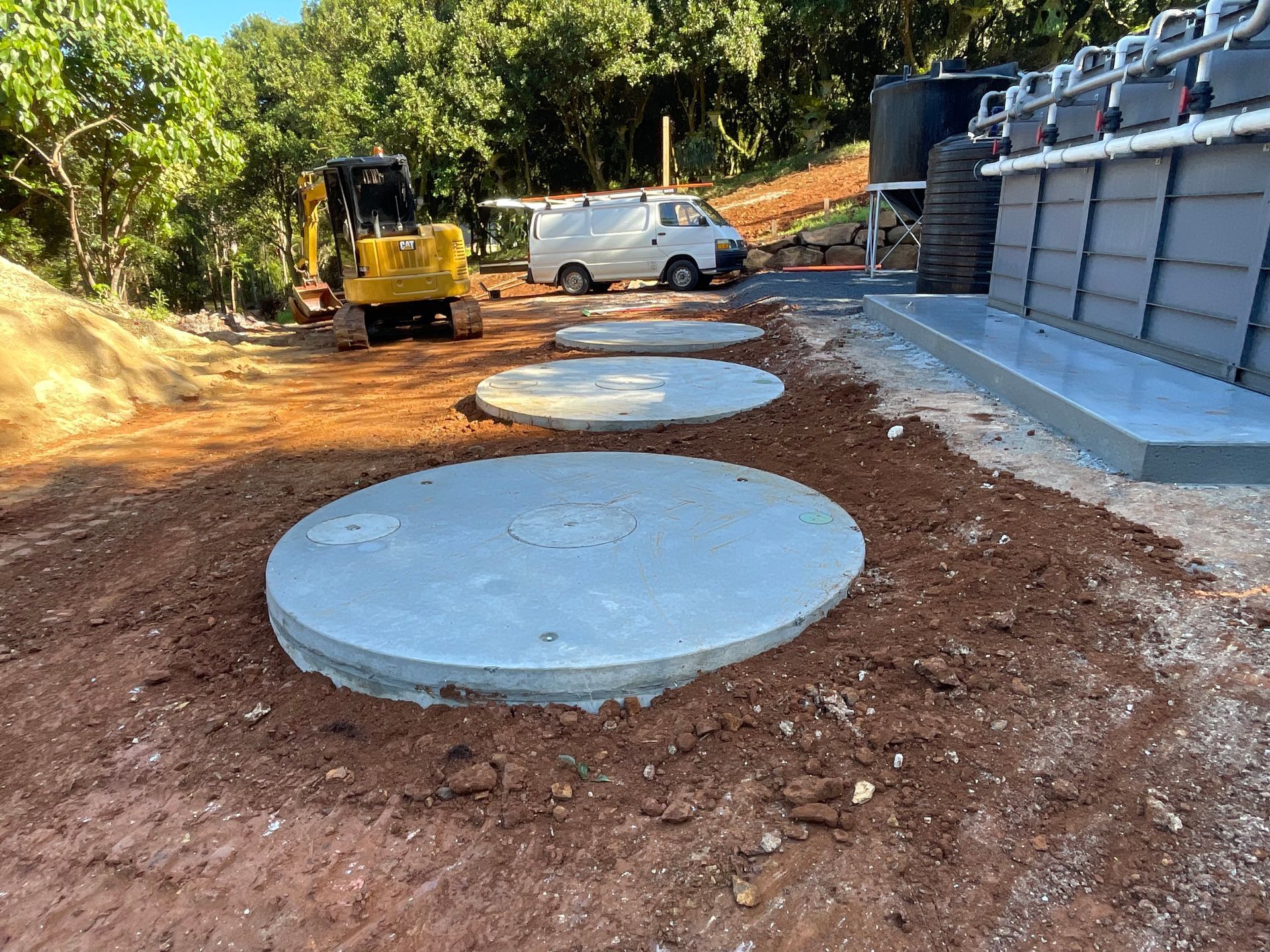 An Excavator Is Working Next To Water Tanks — East Point Earthmoving in Byron Bay, NSW