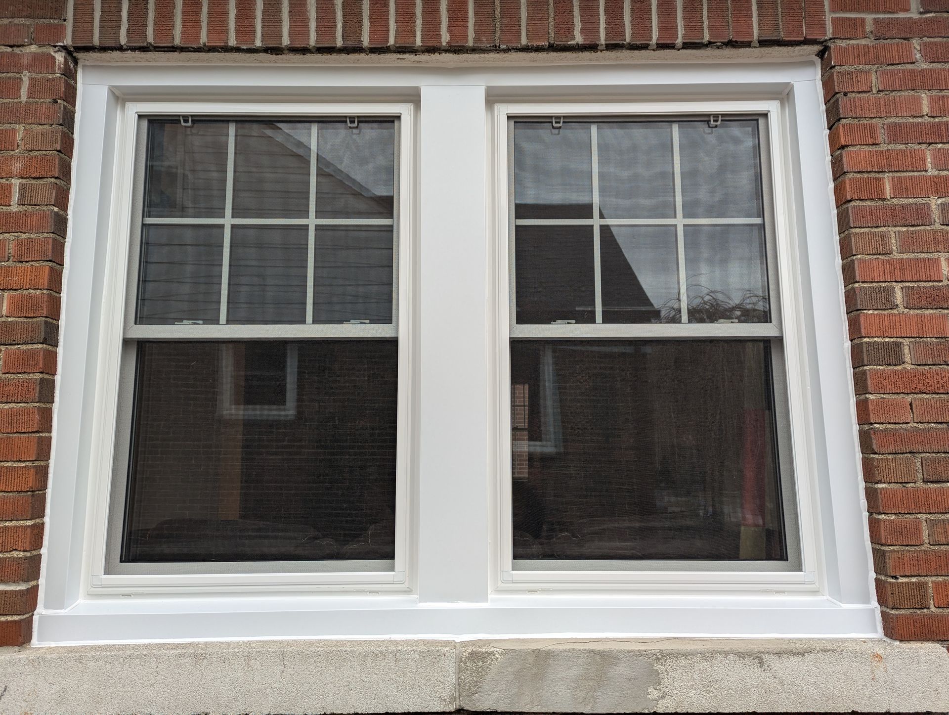 A white, dual-pane, double-hung window with a grid pattern set into a red brick wall above a stone sill.