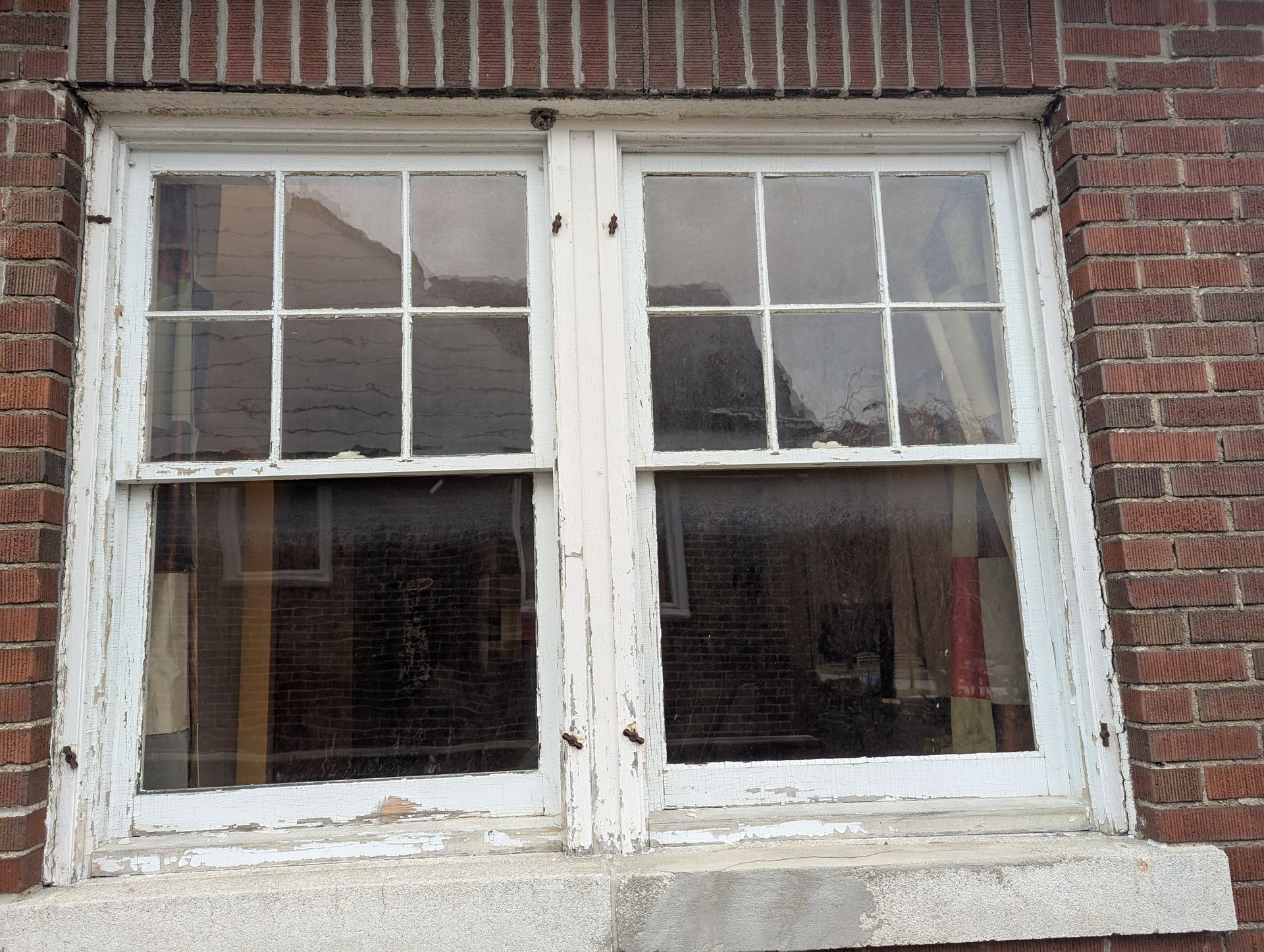 A weathered white-framed, double-hung window with a grid pattern set into a brown brick wall above a concrete sill.