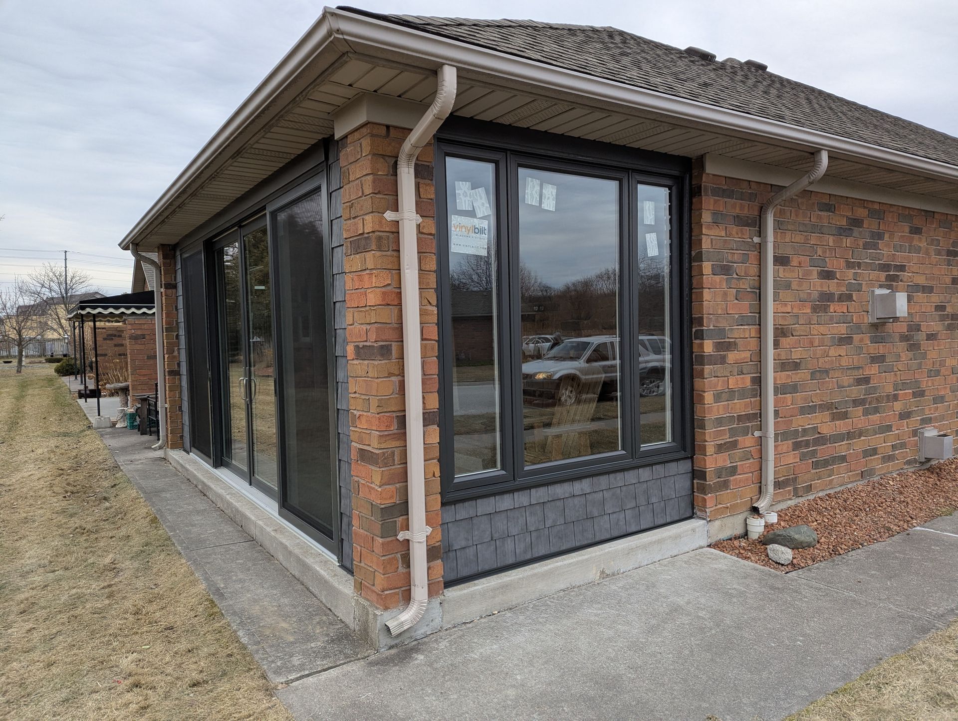 A brick-exterior corner of a house featuring a large, dark-framed window and a glass door on a concrete walkway.