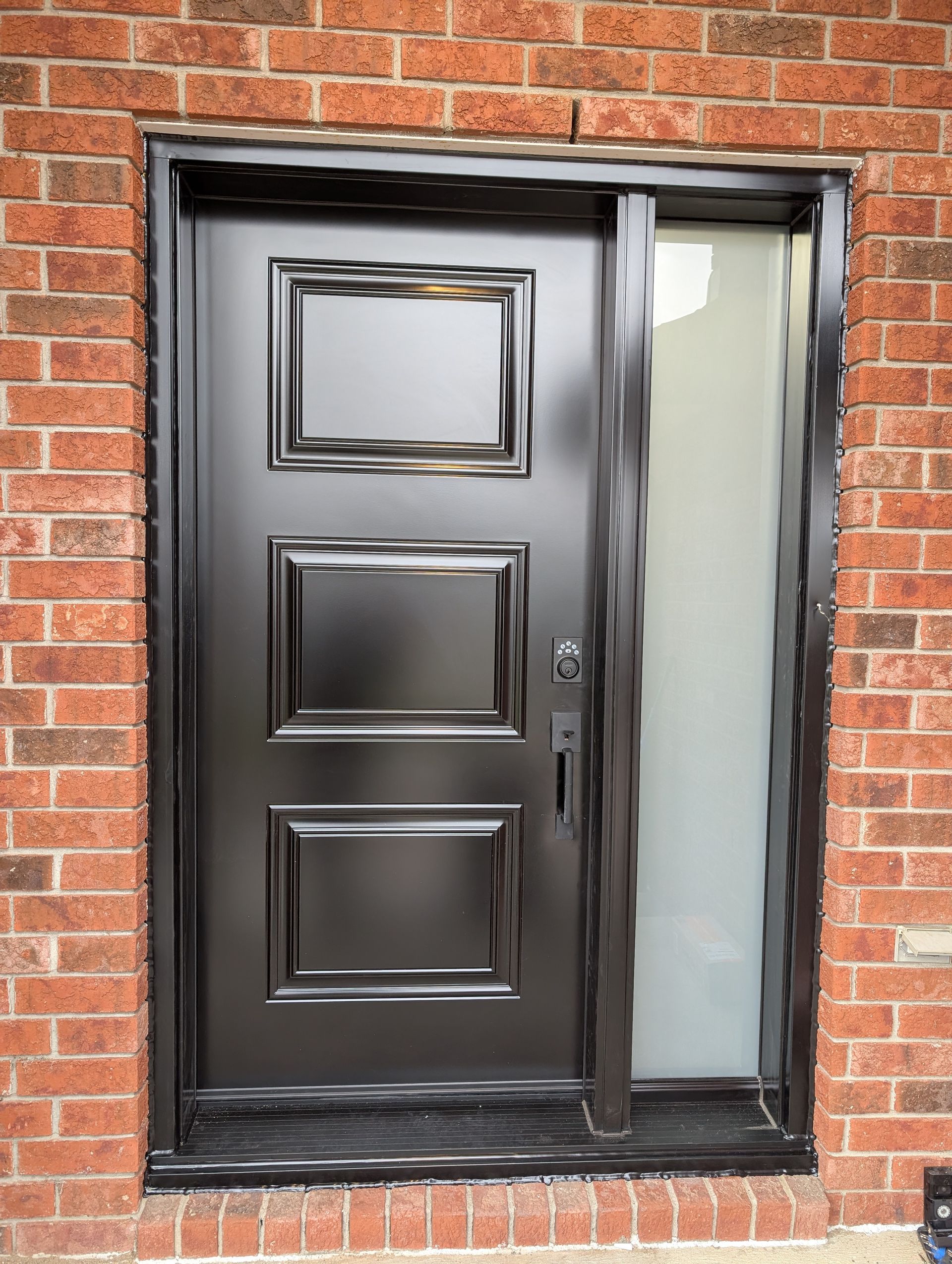 A black front door with three rectangular recessed panels beside a frosted glass sidelight, set in a red brick wall.